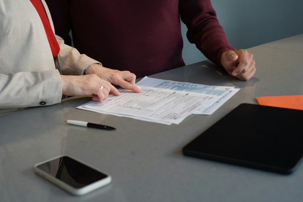 Dos personas junto a una mesa gris revisan unas hojas impresas; una de ellas señala el texto con el dedo